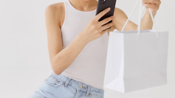 Jonge vrouw bekijkt een webshop op haar telefoon met een winkeltasje in de hand