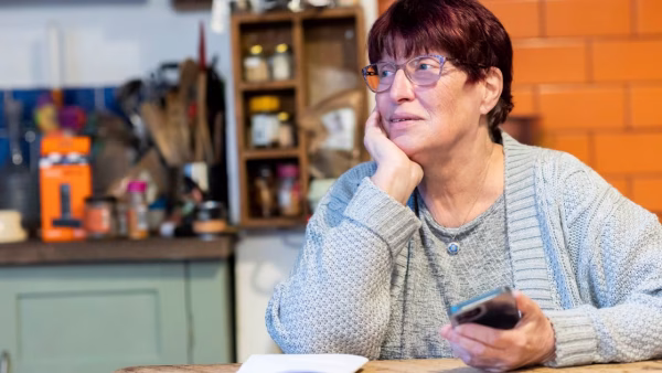 Oudere vrouw kijkt peinzend op met haar telefoon in de hand aan de keukentafel